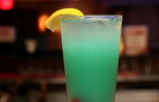 Tall, creamy blue-green cocktail with lemon wedge, ice, and red fruit.