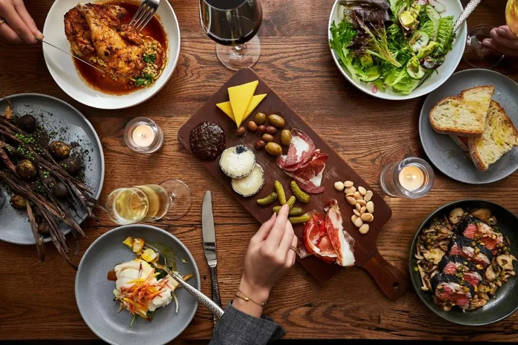 Overhead shot of a dining table with roasted chicken, charcuterie, and hands.
