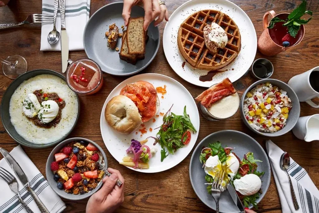 Overhead breakfast and brunch spread on a rustic table with hands.