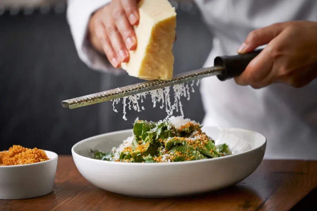 Hands grating Parmesan-like cheese onto a green salad.