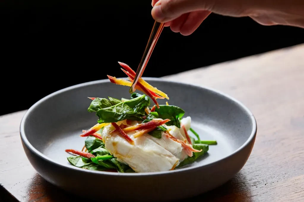 Hand garnishing a gourmet dish of white food and green vegetables.