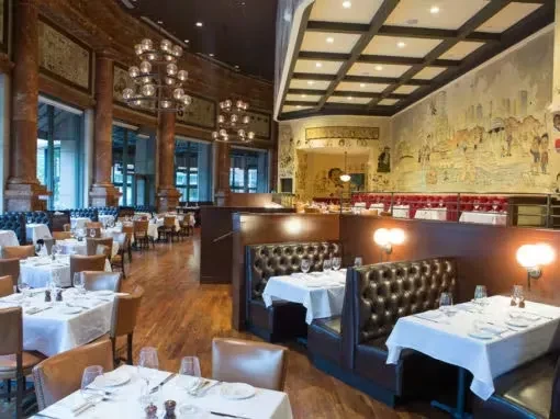 Empty upscale restaurant dining room with set tables, chandeliers, and mural.