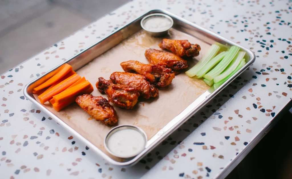 Glazed chicken wings, carrots, celery, and white dip on a metal tray.
