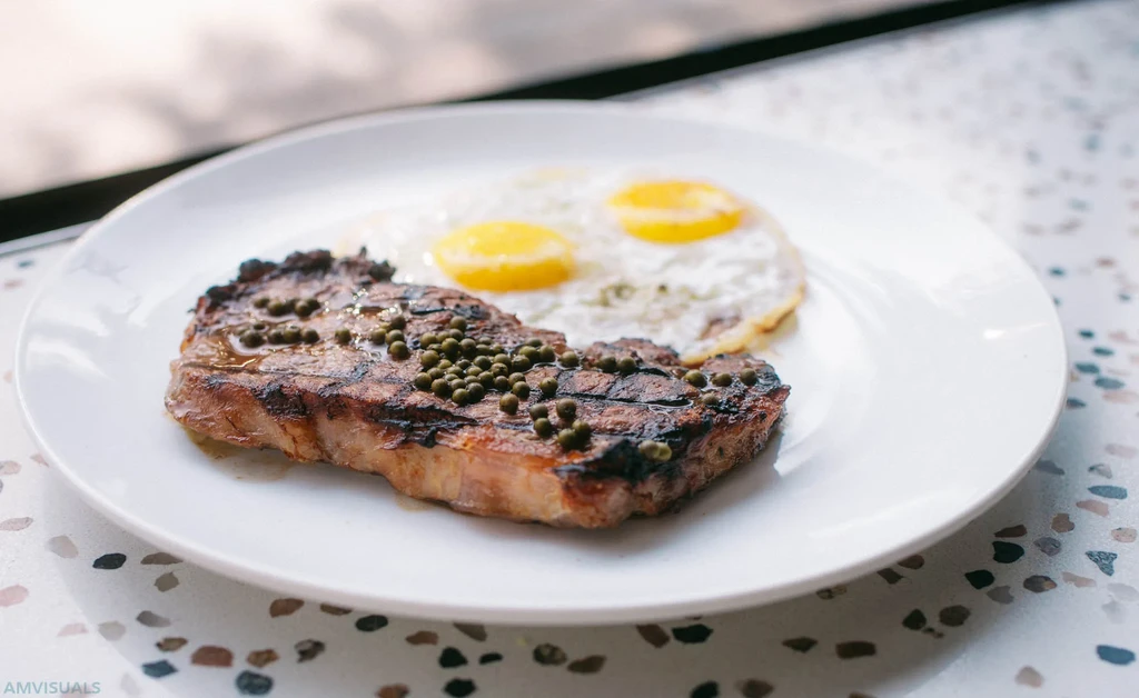 Grilled steak topped with green peppercorns, alongside two sunny-side up eggs.
