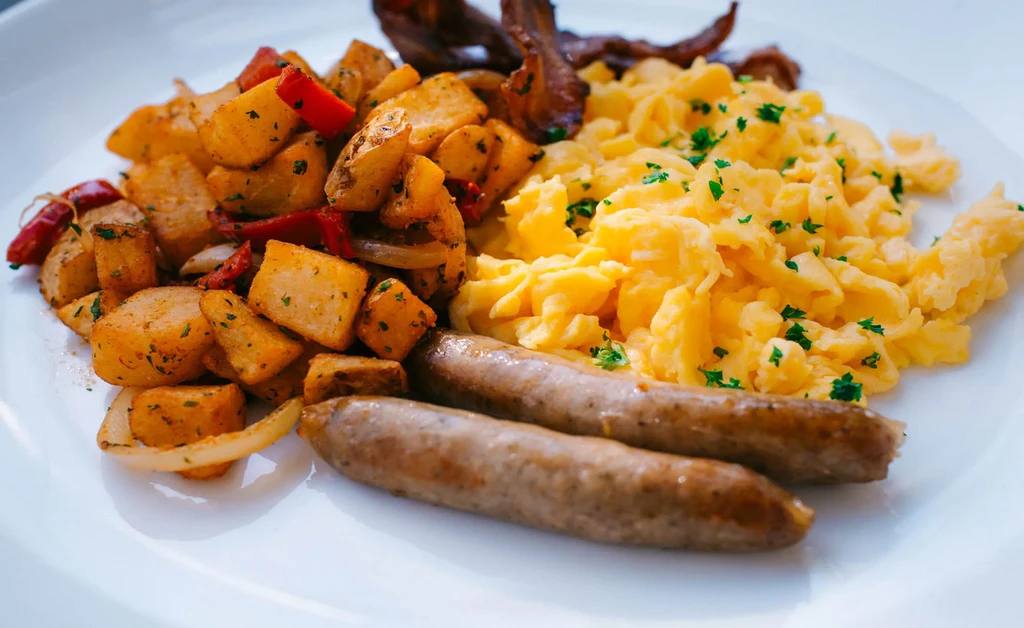 Breakfast plate with scrambled eggs, crispy bacon, sausages, and roasted potatoes.