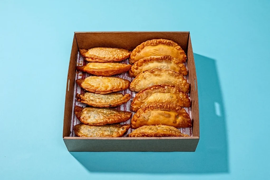 Twelve golden empanadas in a cardboard box on a blue background.