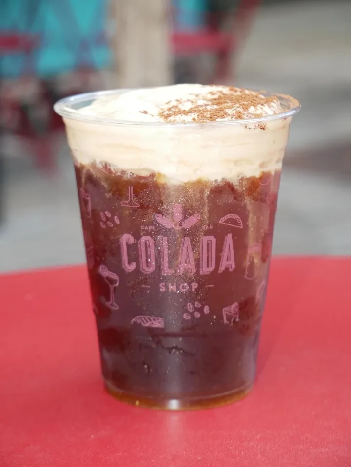 Branded plastic cup with layered cold coffee, creamy foam, and sprinkled powder.