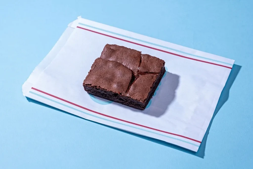 A square chocolate brownie on a striped white wrapper, light blue background.