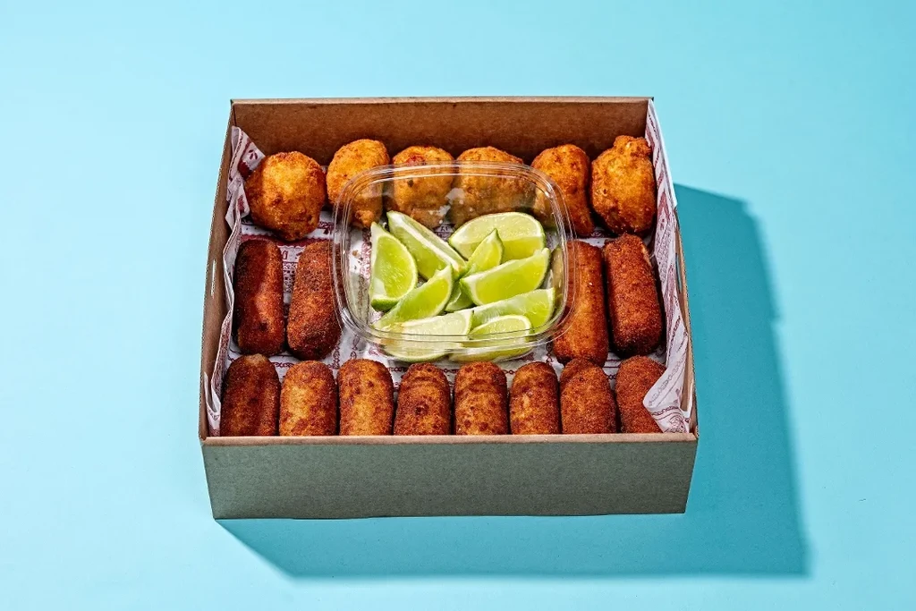 Fried appetizers in a takeout box with lime wedges on a blue background.