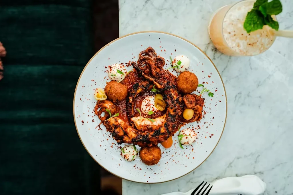 Plated grilled octopus, fried balls, sauces, and a drink on marble.