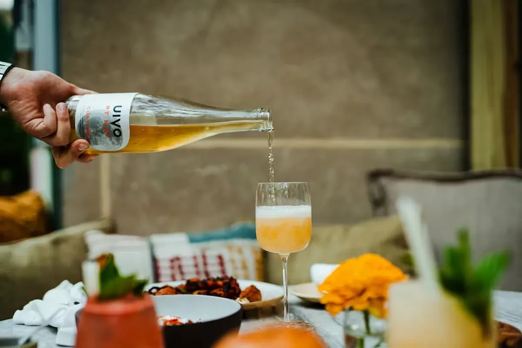 Hand pours golden UIVO liquid into peach drink in a stemmed glass.