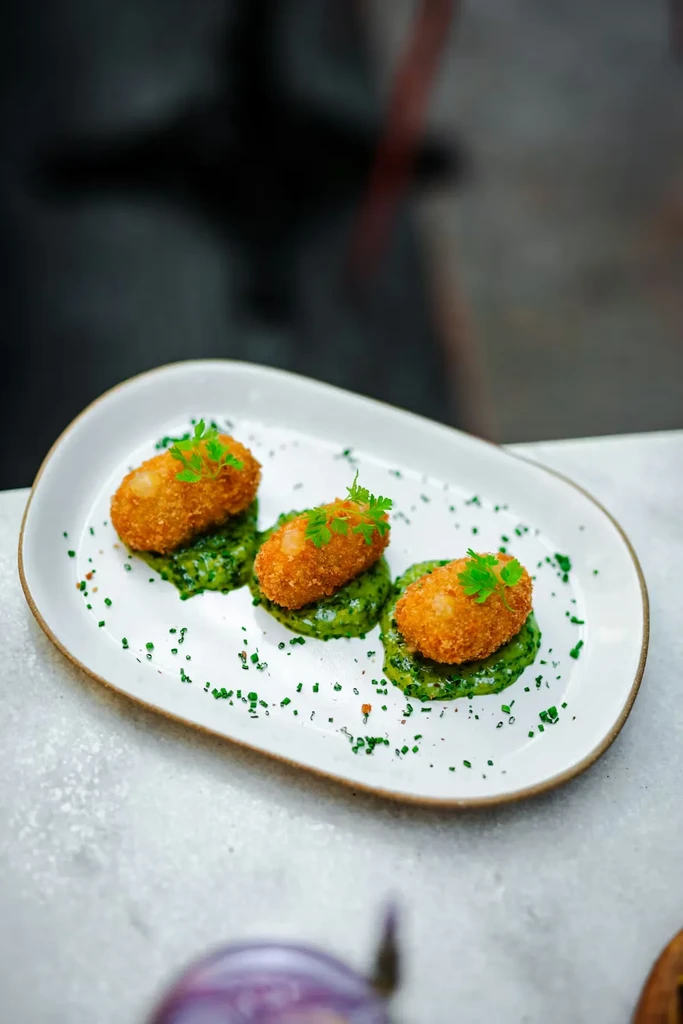 Close-up of three golden-brown croquettes on green sauce and herbs.
