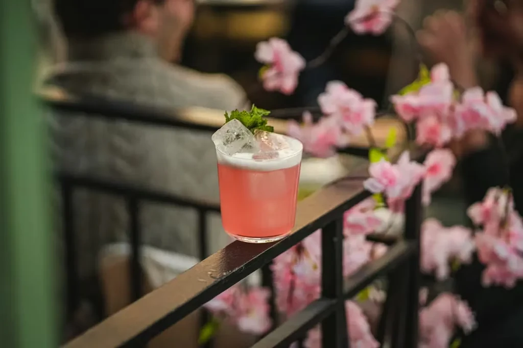 Pink cocktail with foam and mint on dark railing; blurred pink flowers.