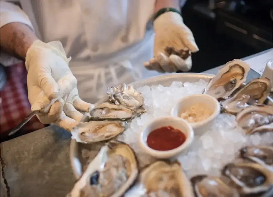 Chef's gloved hands arrange shucked oysters with sauces on ice.