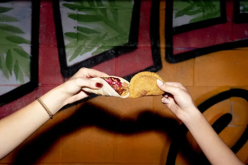 Two hands hold a vibrant taco and an empanada by a graffiti wall.