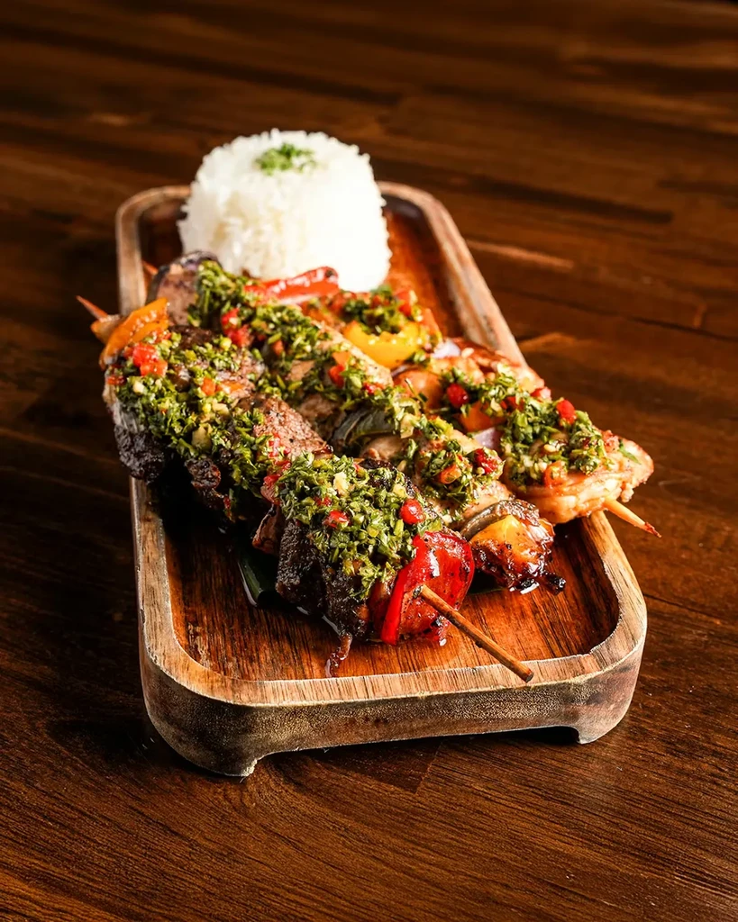 Three grilled meat and vegetable skewers with chimichurri and rice.