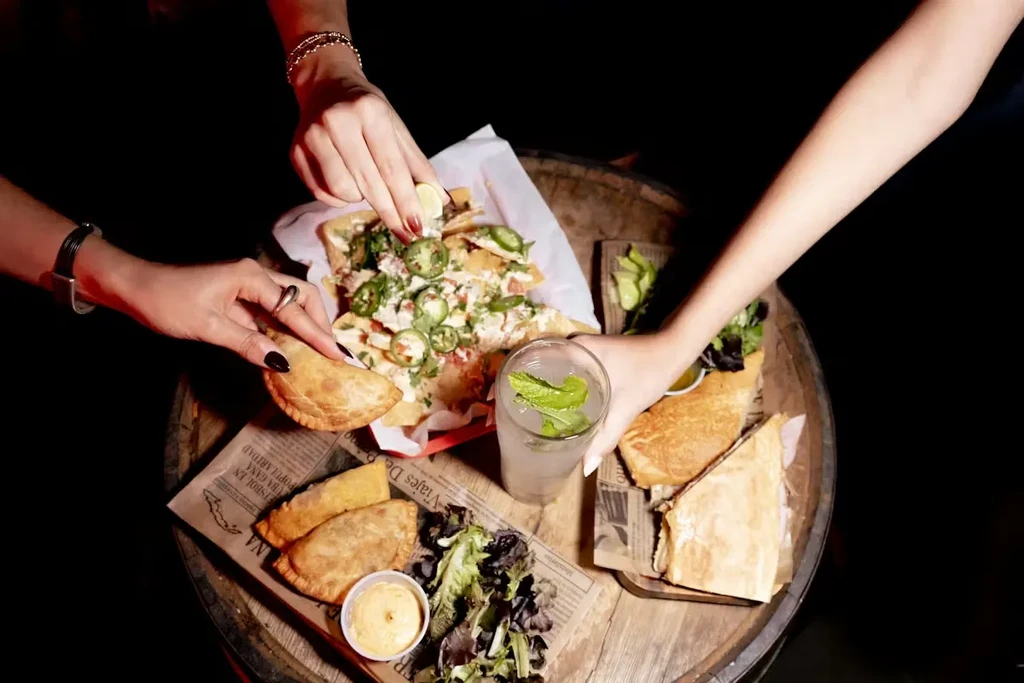 Hands interact with nachos, empanadas, and a drink on a rustic table.