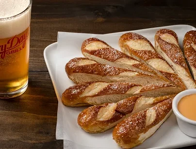 Salted pretzel sticks, orange dipping sauce, and amber beer on wood.