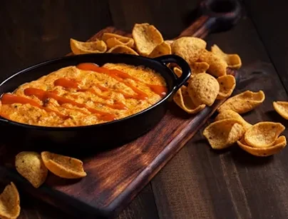Black dish of orange dip with tortilla chips on a wooden board.