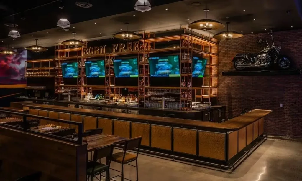 Industrial bar interior with U-shaped counter, liquor shelves, TVs, and motorcycle.