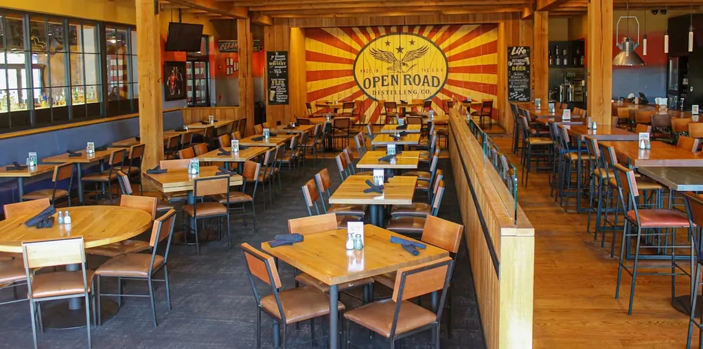 Empty rustic industrial restaurant with dining tables, bar, and distillery sign.