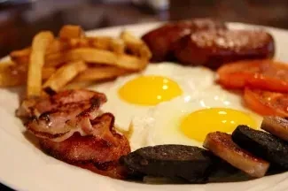 Breakfast platter with fried eggs, bacon, sausages, fries, tomatoes, and black pudding.