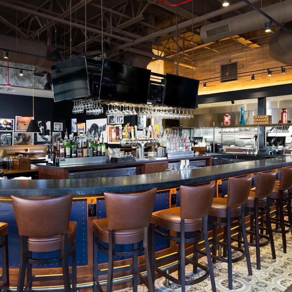 Modern restaurant bar with empty leather stools, bottles, and TVs.