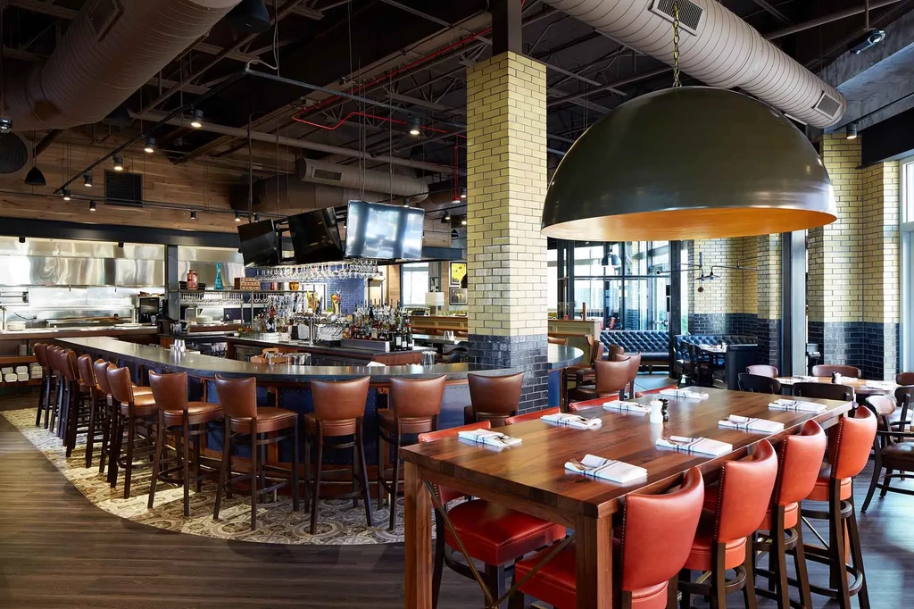 Modern industrial-chic restaurant interior with a bar, communal table, and booths.