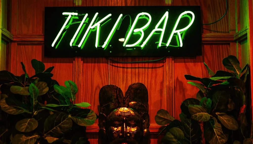 Green 'TIKI-BAR' neon sign above a tiki mask and tropical plants.