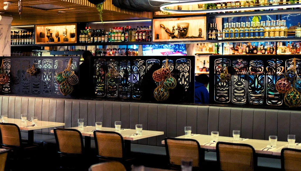 Restaurant interior featuring dining tables, booths, and a brightly lit tiki bar.