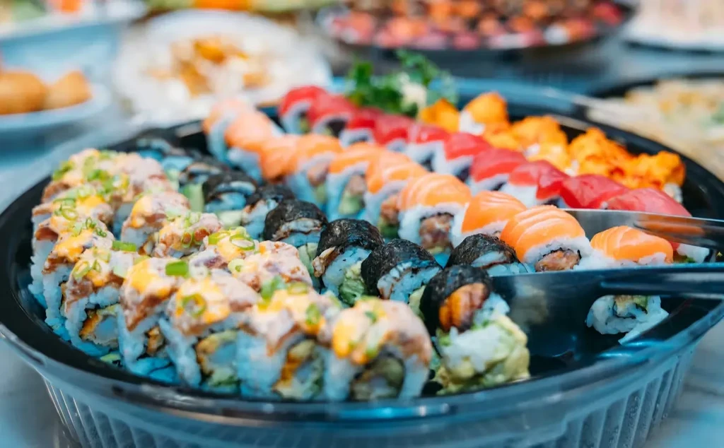 Assorted sushi rolls in a black serving tray with tongs.
