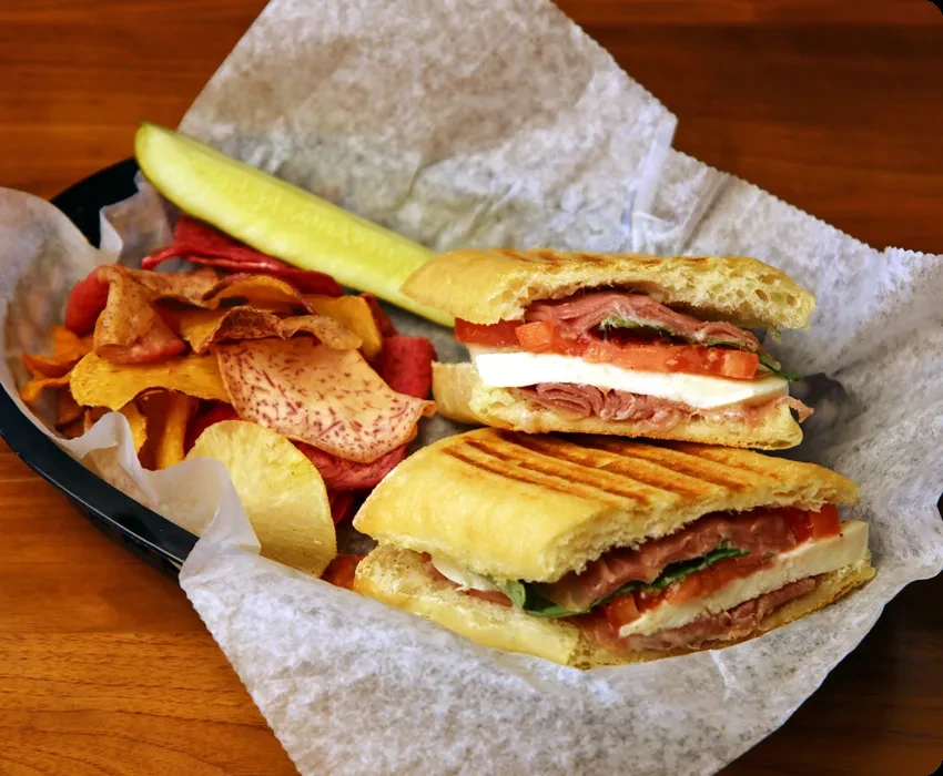 Grilled panini sandwich with deli meat, mozzarella, colorful chips, and pickle.