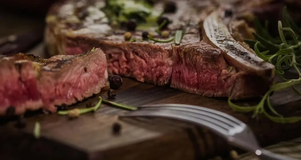 Sliced medium-rare steak, rosemary, peppercorns on a dark wooden board.