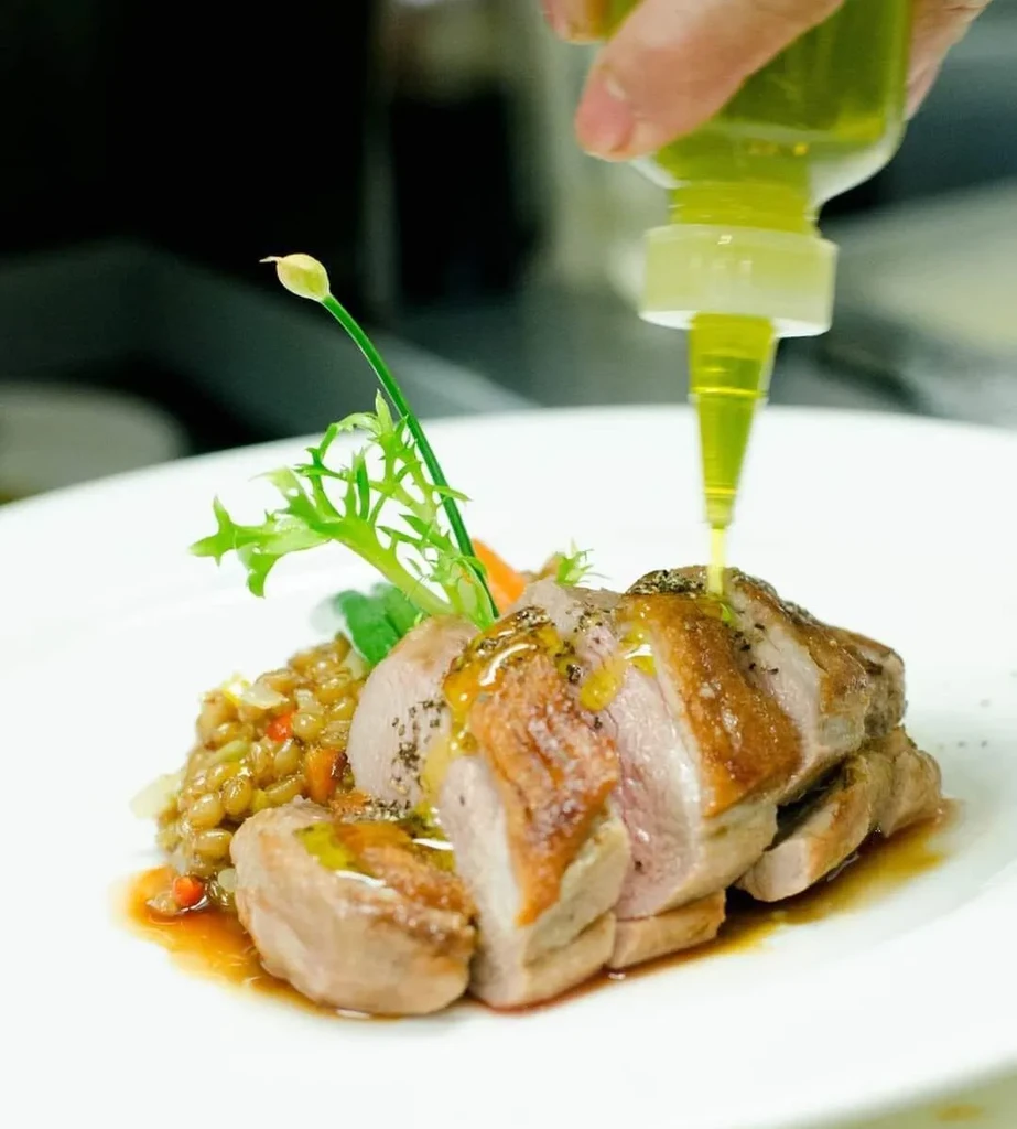 Hand drizzles sauce onto sliced duck, lentils, and fresh greens.