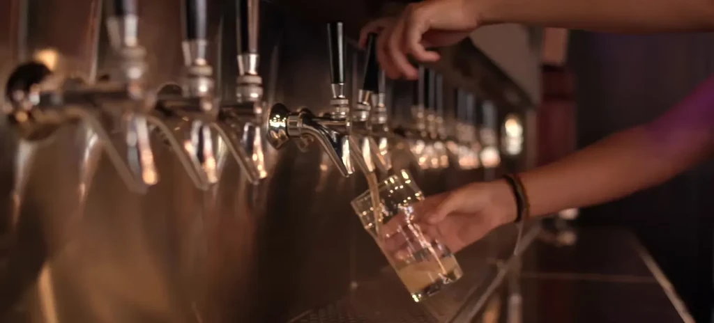 Hands pouring golden liquid from a tap into a glass at a bar.