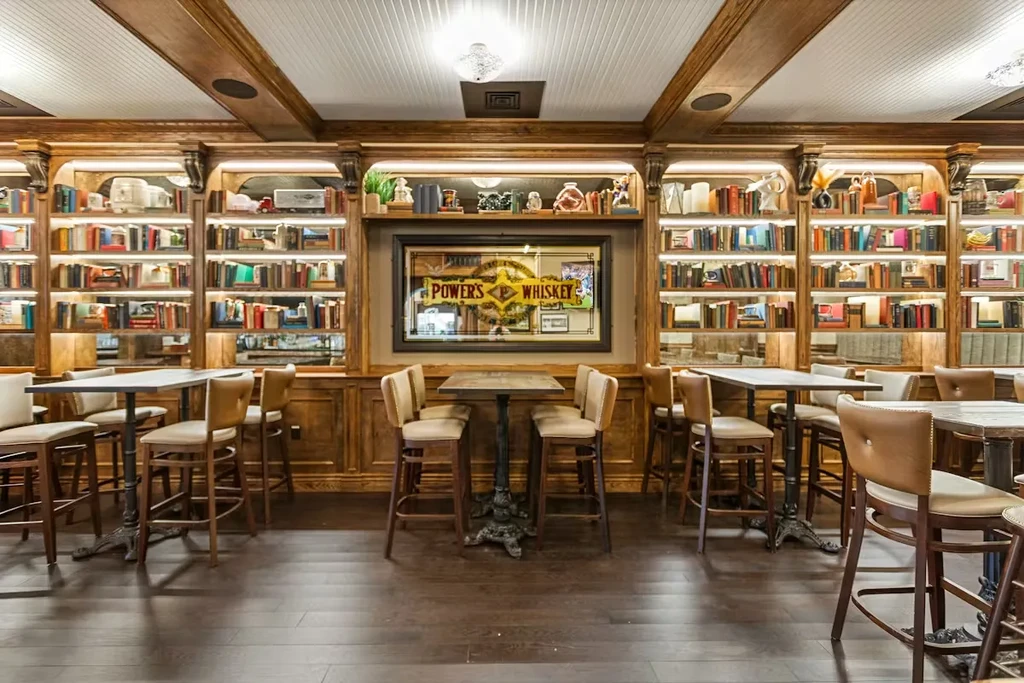 Cozy bar interior with bookshelves, high tables, and "POWERS WHISKEY" mirror.