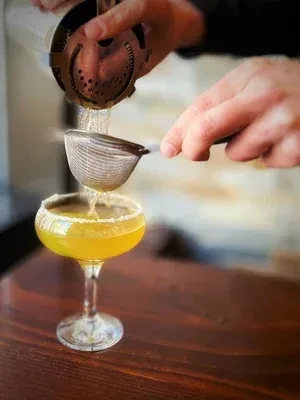 Close-up of hands double-straining a cocktail into a rimmed coupe glass.