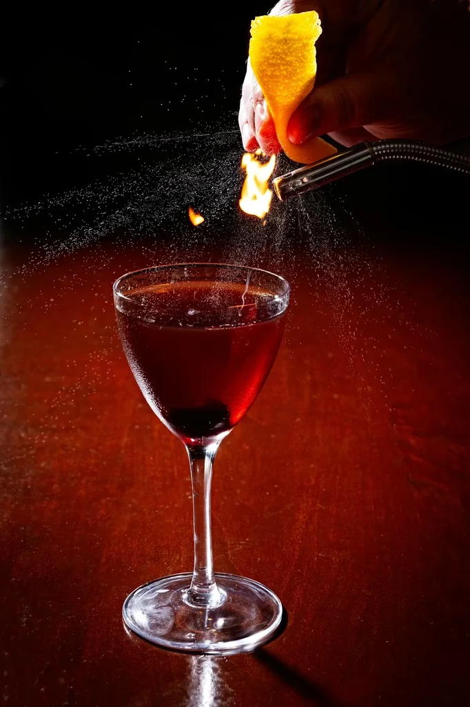 Hand expresses flaming orange oil over a dark red cocktail.