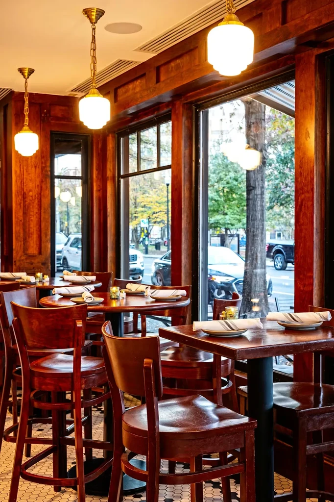 Warmly lit restaurant with wooden tables, dark paneling, and street views.