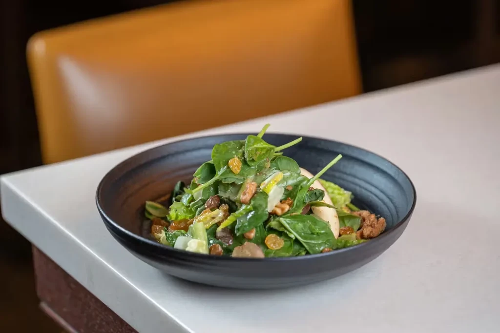 Salad with greens, golden raisins, nuts, and fruit in dark bowl.