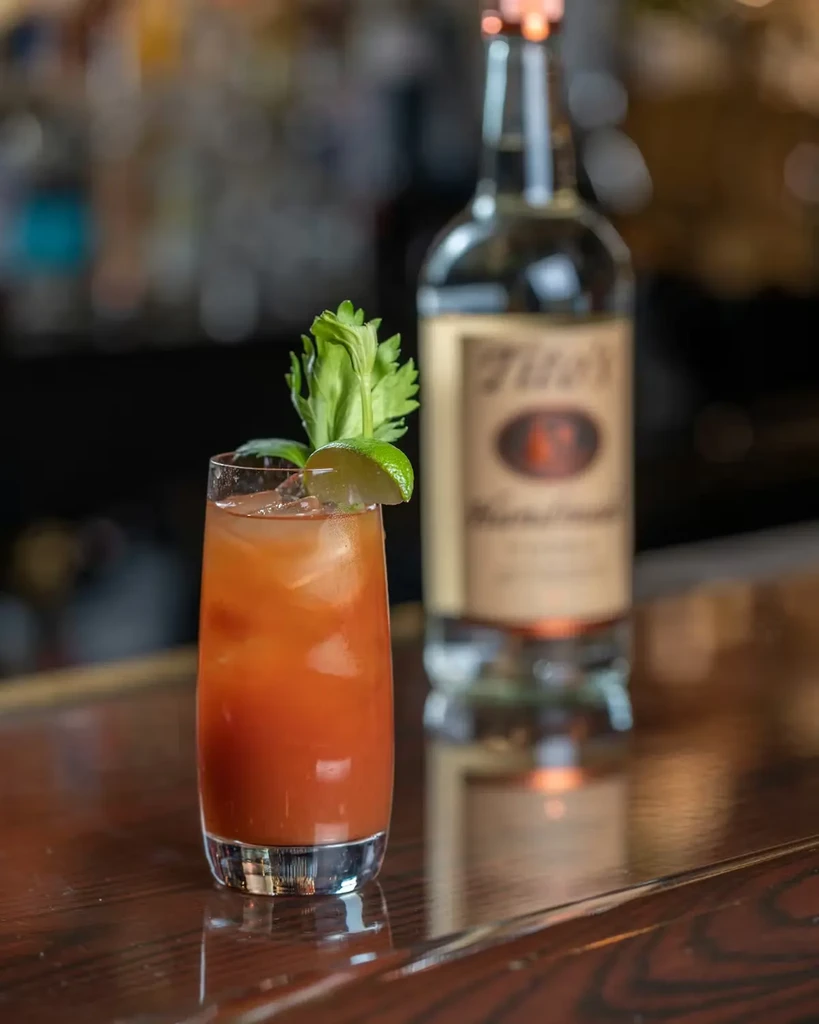 Orange-red cocktail with ice, celery, and lime on a wooden bar.