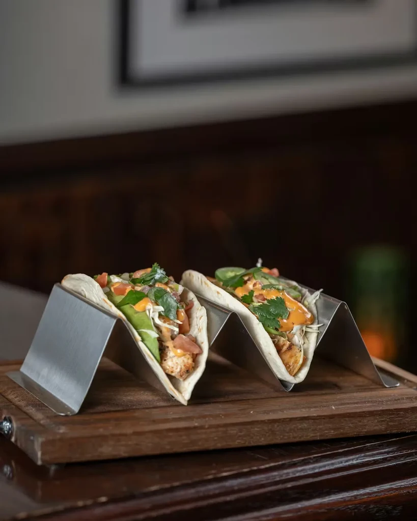 Two generously filled soft tacos in metal holders on a wooden board.