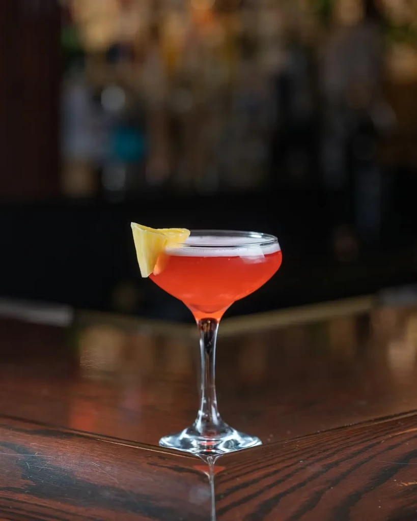 Coral cocktail in coupe glass with pineapple wedge on dark bar.