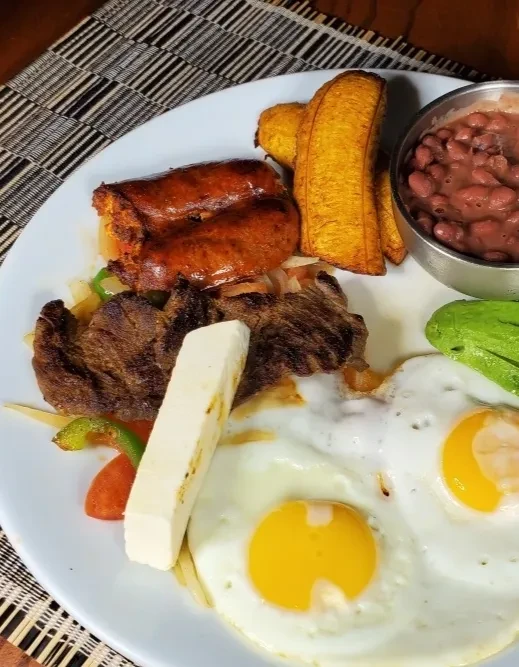 Hearty plate of steak, sausages, eggs, plantains, beans, cheese, avocado.