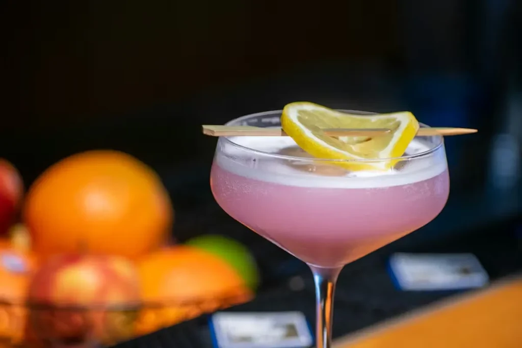Pink cocktail, frothy head, lemon on skewer, blurred fruit background.