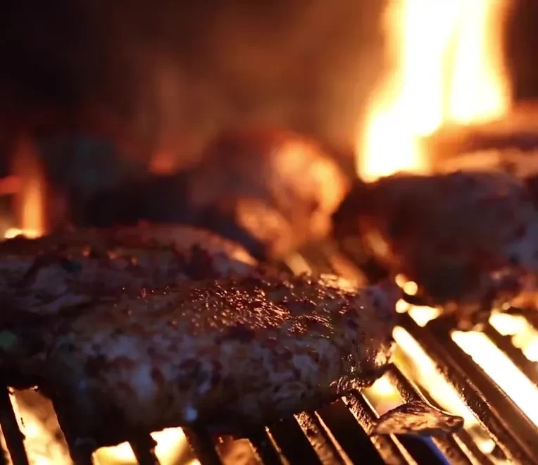 Charred, seasoned meat sizzling on glowing grill over strong open flames.