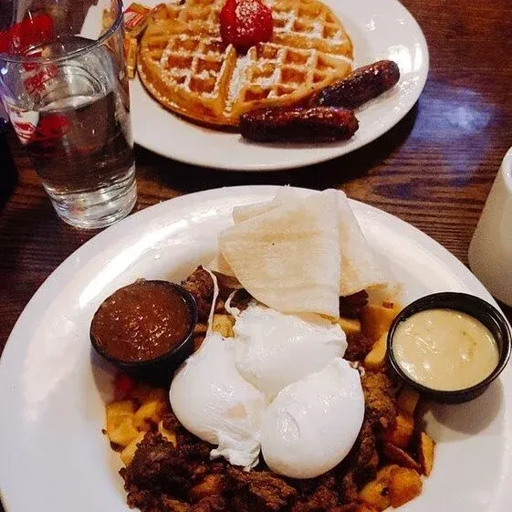Two plates of breakfast and brunch: waffle, hash, eggs, sausages.