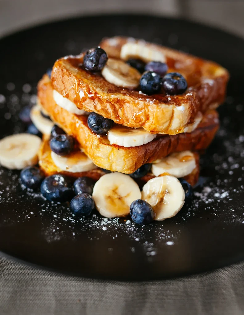 Stack of golden French toast with bananas, blueberries, syrup, powdered sugar.