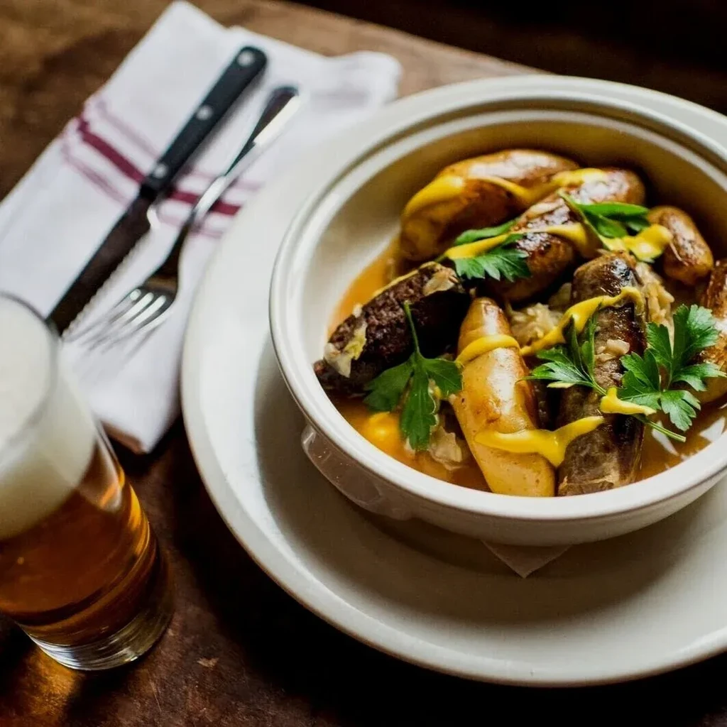Bowl of sausages and potatoes with mustard, beer, and cutlery.
