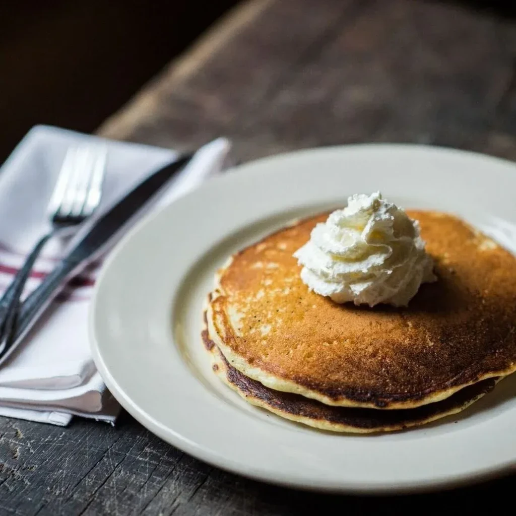Stack of pancakes with whipped cream and cutlery on a dark wood table.